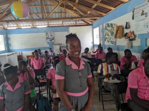 Smiling Haitian teenage girl wears a gray and pink uniform and stands in a classroom with other students