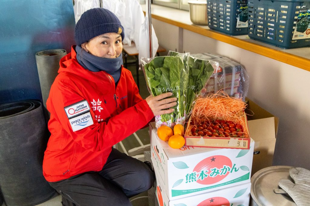 Japanese woman wearing winter hat, scarf, and red ARROWS jacket kneels next to boxes of produce with oranges, lettuce, and tomatoes