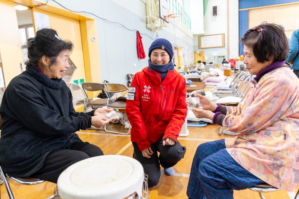 Two smiling Japanese women sit on opposite sides in an elementary school gym holding a plastic wrapped food while a Japanese woman in a red ARROWS jacket sits between them