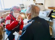 Doctor wearing a red ARROWS uniform and white face mask looks into the mouth of an elderly man with a flashlight