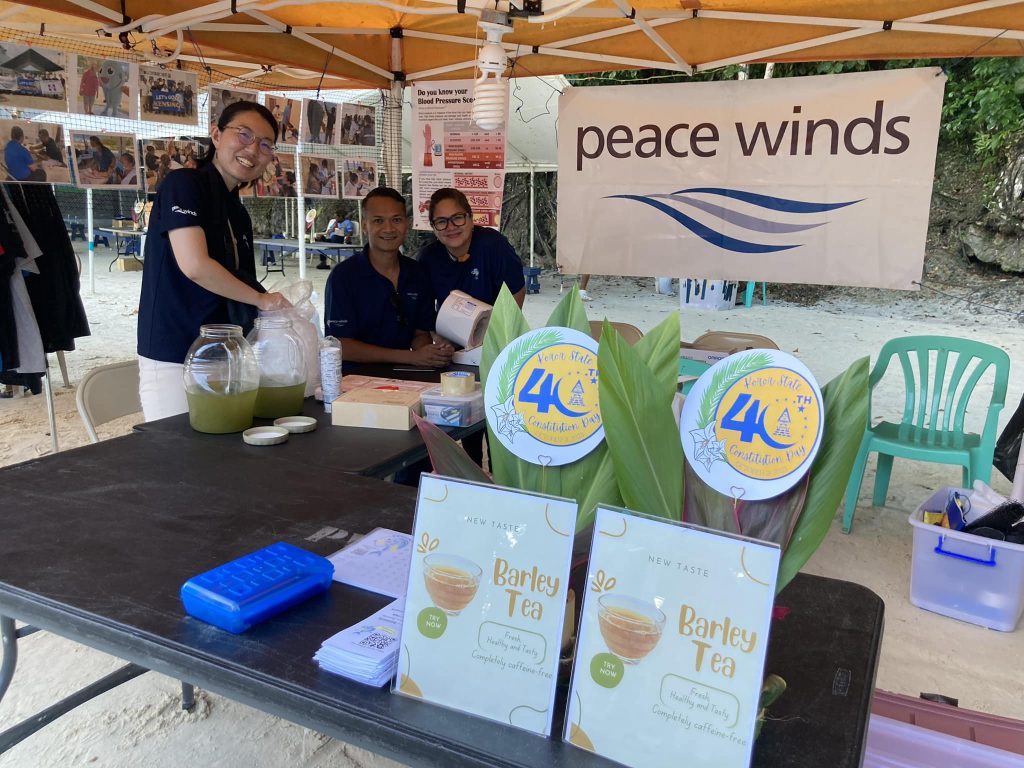 3 Peace Winds Palau staff smile next to a booth/table with informational signs