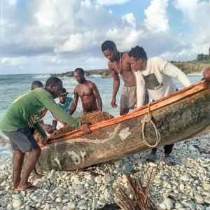 6 Haitian men drag a small wooden boat on a rocky shore next to blue water