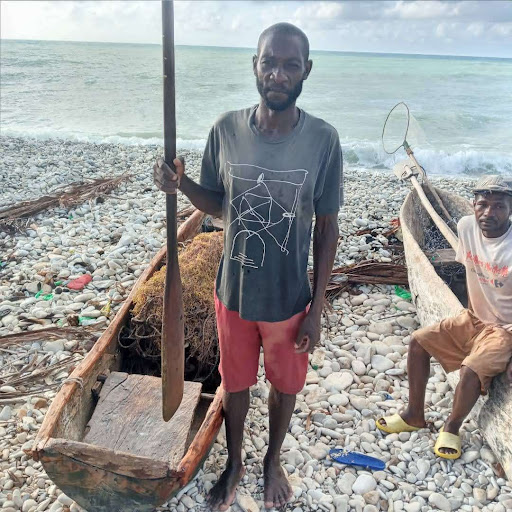 Haitian man wearing a t-shirt and shorts holds a wooden oar and stands next to a small wooden boat on a rocky shore with blue water and sky in the background