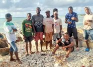 Group of Haitian men wearing t-shirts and shorts stands on a rocky beach with blue water and sky in the background