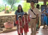Japanese man wearing white Peace Winds polo shirt stands with a woman in a blue t shirt and two young girls wearing red traditional outfits