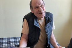 Elderly man sits on a bed with a blue plaid blanket. He is wearing an off-white t-shirt and a blue vest