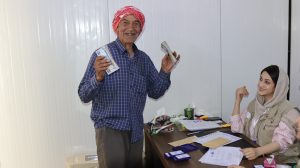 Smiling Syrian man wearing a red turban and blue button-down shirt holds cash in both hands