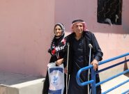 An elderly Syrian man and woman stand on a concrete wheelchair ramp with a blue handrail
