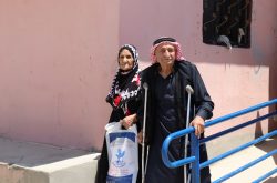 An elderly Syrian man and woman stand on a concrete wheelchair ramp with a blue handrail