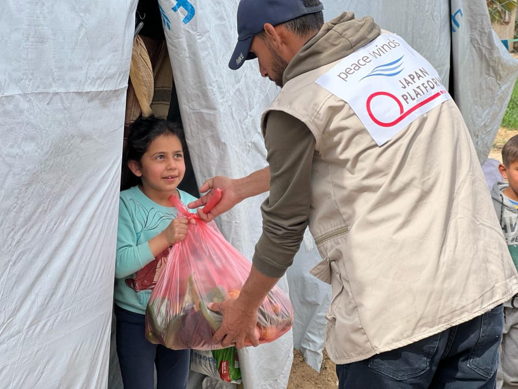 Man wearing a canvas Peace Winds vest hands a bag full of food to a young girl