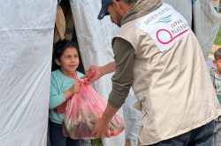 Man wearing a canvas Peace Winds vest hands a bag full of food to a young girl
