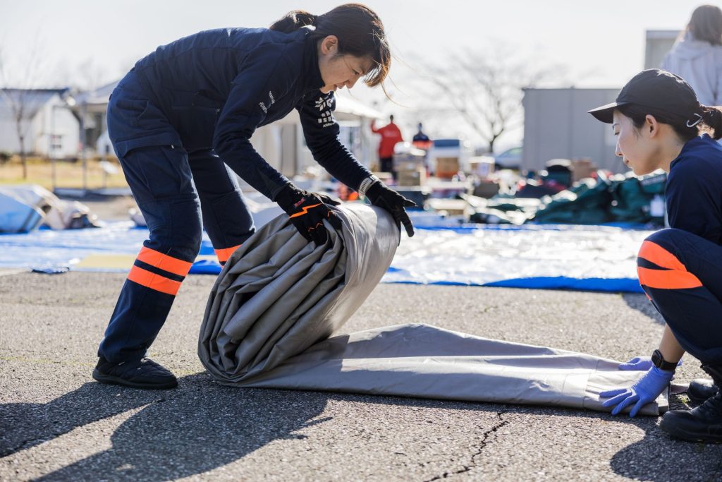 Two women in ARROWS uniforms unroll a plastic tent