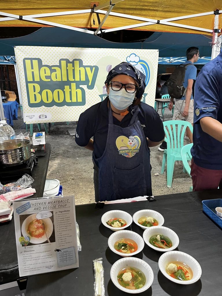Woman wearing a mask and glasses stands behind a table with small bowls of soup in front of a banner with yellow text that says "healthy booth"