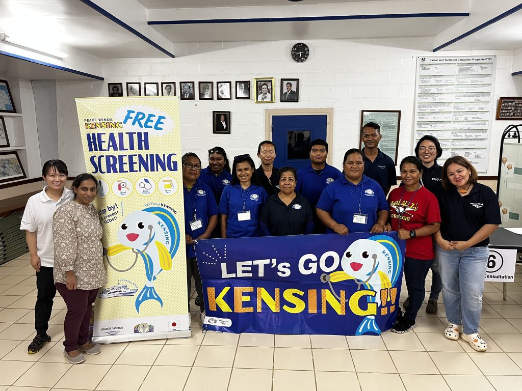 Group of Peace Winds staff members wearing dark blue polo shirts poses next to a yellow banner that says "free health screening" and a blue banner with a cartoon fish that says "let's go kensing"
