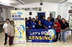 Group of Peace Winds staff members wearing dark blue polo shirts poses next to a yellow banner that says "free health screening" and a blue banner with a cartoon fish that says "let's go kensing"