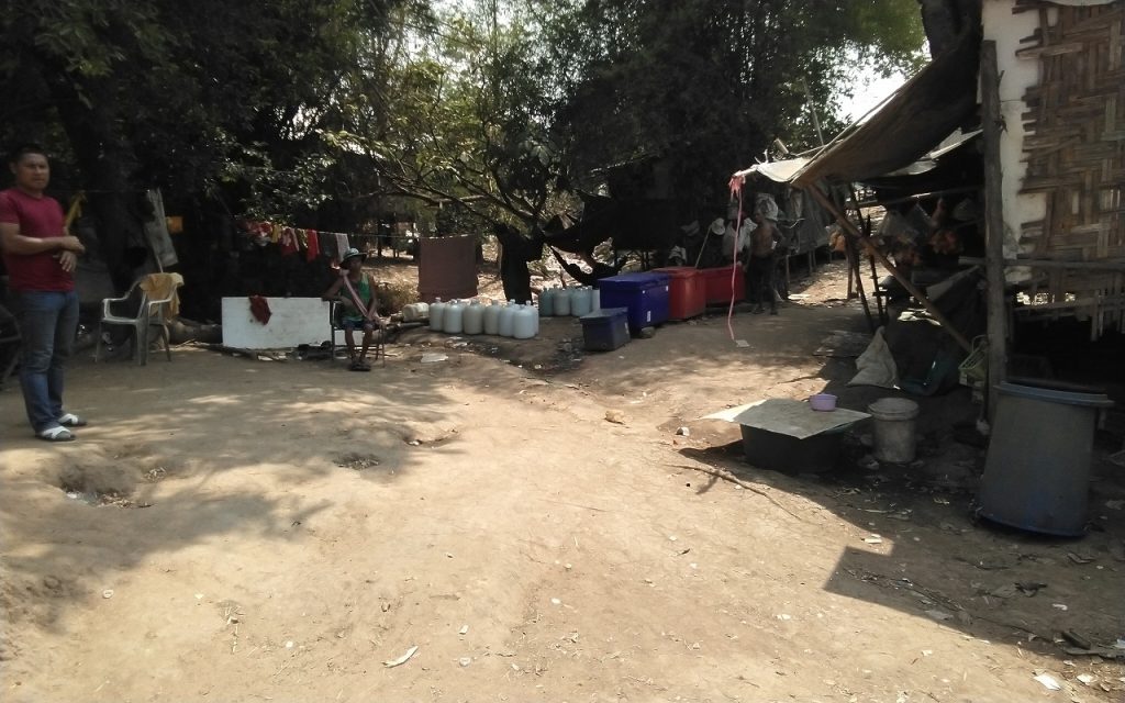 View from a dirt ground/road of a simple shelter with water jugs on the ground outside it