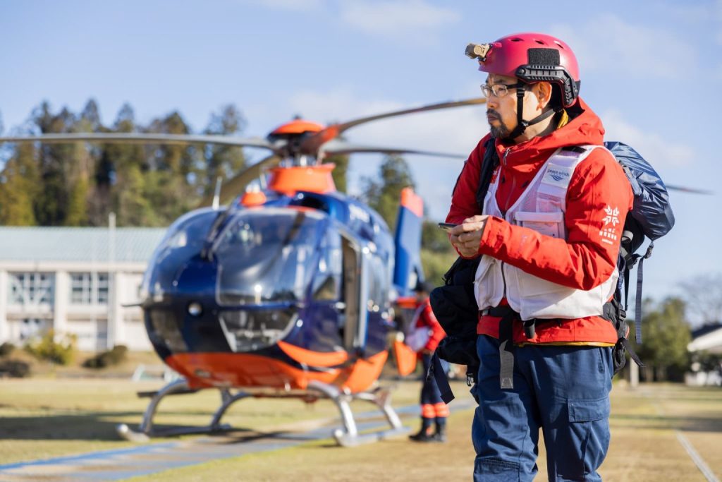 man wearing a red search-and-rescue uniform with white vest walks toward the camera and there is an orange helicopter in the background