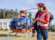 man wearing a red search-and-rescue uniform with white vest walks toward the camera and there is an orange helicopter in the background