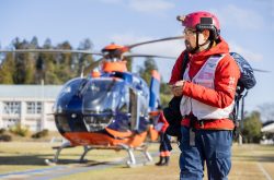 man wearing a red search-and-rescue uniform with white vest walks toward the camera and there is an orange helicopter in the background