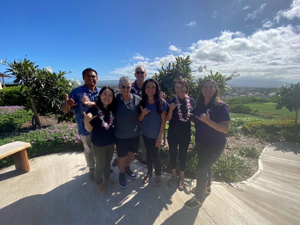 seven people wearing blue shirts and pink flower leis stand outside in a group while smiling for the picture