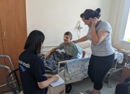 Boy with an eyepatch sits up in a hospital bed while a woman stands and looks at him with a hand on her back and the boy talks to a woman sitting in a chair with a Peace Winds logo t shirt