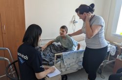 Boy with an eyepatch sits up in a hospital bed while a woman stands and looks at him with a hand on her back and the boy talks to a woman sitting in a chair with a Peace Winds logo t shirt