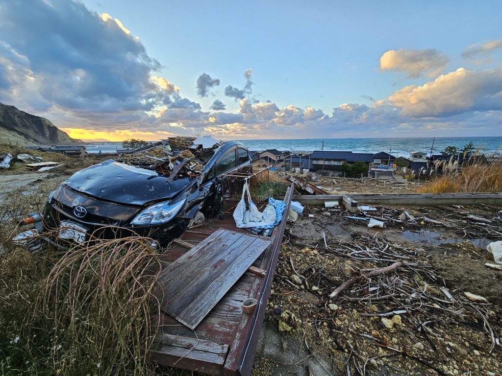 Landscape with debris and a destroyed car in the foreground and a sunset, ocean, and a village with destroyed homes in the background