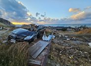 Landscape with debris and a destroyed car in the foreground and a sunset, ocean, and a village with destroyed homes in the background