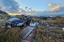 Landscape with debris and a destroyed car in the foreground and a sunset, ocean, and a village with destroyed homes in the background