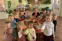 11 ukrainian children and one teacher face the camera while standing around a craft table in a classroom. They are holding out a craft they made