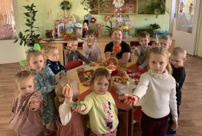 11 ukrainian children and one teacher face the camera while standing around a craft table in a classroom. They are holding out a craft they made