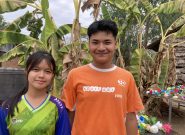 two smiling burmese teenagers, a boy and girl, stand in front of palm trees