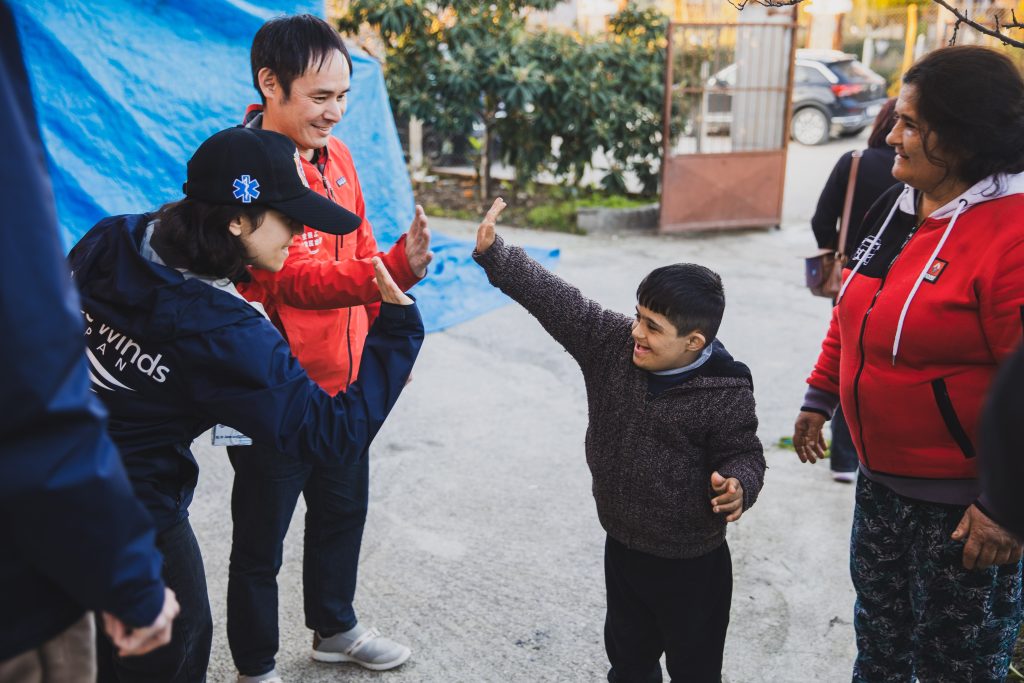 A Japanese woman and man smile and high-five a smiling Turkish boy. A Turkish woman is standing next to them, and they are all standing outside on a concrete ground in front of trees, a metal gate, and a blue tarp-covered structure.