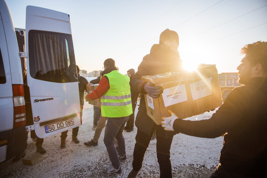 In the foreground, a man in winter clothing passes a cardboard box with the Peace Winds logo to another man. There are other people passing boxes to one another next to a large white van in the background, and there is a sunlight flare over the first man's shoulder.