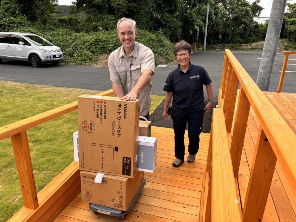 A white man and a Japanese woman stand next to each other on a wooden ramp smiling at the camera. The man is pushing a dolly with cardboard boxes of small appliances.