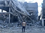 a man stands with his back to the camera wearing a tan vest with the Peace Winds logo on the back. In front of him is the rubble of a destroyed building.