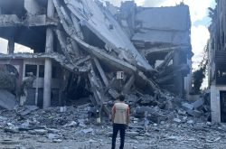 a man stands with his back to the camera wearing a tan vest with the Peace Winds logo on the back. In front of him is the rubble of a destroyed building.
