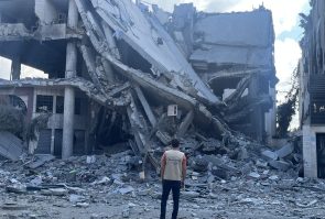 a man stands with his back to the camera wearing a tan vest with the Peace Winds logo on the back. In front of him is the rubble of a destroyed building.