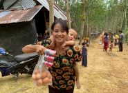 A smiling young Burmese woman wearing a black shirt with sunflowers has a baby on her back and holds up a bag of eggs and canned fish. She is standing outside on a dirt road, and there are people walking away in the background, trees, and a metal building