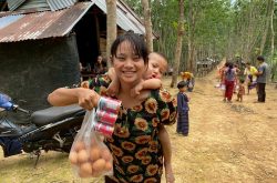 A smiling young Burmese woman wearing a black shirt with sunflowers has a baby on her back and holds up a bag of eggs and canned fish. She is standing outside on a dirt road, and there are people walking away in the background, trees, and a metal building