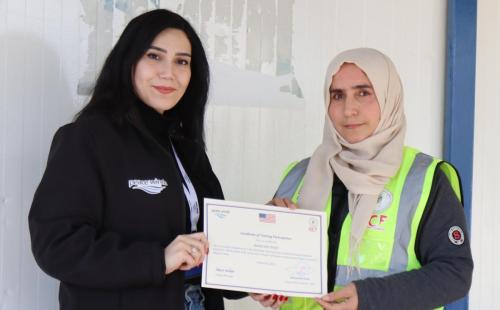 A woman wearing a black jacket with Peace Winds logo and a woman in a yellow safety vest and white hijab stand together holding a certificate