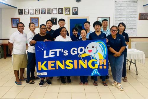 A dozen people pose with a dark blue banner that says "let's go kensing" and has a blue cartoon surgeonfish
