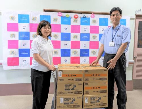 Two Japanese staff members stand with a cart full of cardboard boxes in front of a pink and blue banner