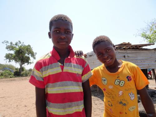 Two Mozambican boys, young teenagers, smile in the foreground. One is wearing a yellow shirt and one is wearing a red striped shirt. They are standing outside in a background of trees and a dirt ground.