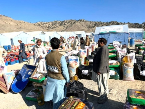 Outdoor mountainous landscape with light blue tents, piles of food packages, and several people gathered in the camp